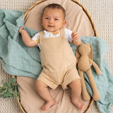 A baby lying on a woven mat with a beige cushion, wearing beige overalls over a white shirt, with a mint green blanket and a tan stuffed bunny toy.
