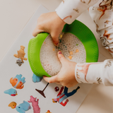 Child playing with a green toy filled with colorful beads on a white surface with stickers.