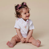 A smiling baby girl sitting on a beige studio background, wearing a white short-sleeve bodysuit and a white bib with a pink and yellow ice cream cone print. She has her hair in two buns with pink bows.