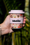 Hand holding a pink ice cream-themed cup with 'bbeeño' branding against a blurred green leafy background