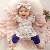 A baby lying on a pink ruffled blanket, wearing a white floral dress with flutter sleeves, a matching bow headband, and purple leggings.