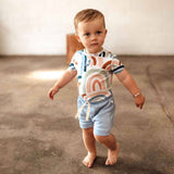 Baby standing on a concrete floor wearing a colorful outfit with a rainbow design.