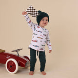 Child wearing a car-themed shirt and black pants, holding a checkered flag next to a red toy car on a beige background