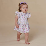 A toddler girl wearing a white short-sleeve dress with a colorful floral print, standing against a solid beige background. She has a purple hair bow in her hair.