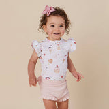 A toddler girl with curly brown hair wearing a white short-sleeve top with a colorful ice cream print and light pink ruffled shorts, standing against a solid beige background.