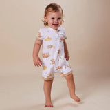 A smiling toddler stands barefoot against a plain beige studio background, wearing a white short-sleeve sleep sack or romper with a light tan and yellow teddy bear print and beige trim.