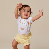 A baby wearing a white short-sleeve bodysuit with a teddy bear print and pale yellow ruffled shorts, standing on a plain beige studio background with arms raised.