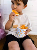 Child eating an orange slice while holding a cup with more oranges, sitting on a wooden floor.