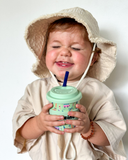 Child wearing a beige sun hat holding a mint green cup with a straw against a plain background