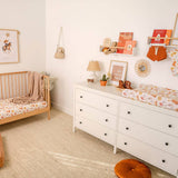 A styled nursery room featuring a white wooden dresser with a changing pad, a light wood crib, and warm neutral decor on a beige rug.