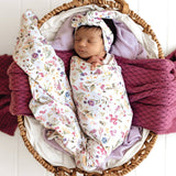 A sleeping newborn baby swaddled in a white floral blanket with matching knotted headband, resting inside a wicker basket with purple knit blankets and a white fabric lining.