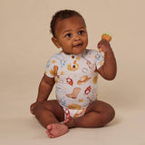 A baby sitting on a beige studio floor wearing a white short-sleeve bodysuit with a colorful western print, holding a small orange and yellow rattle.