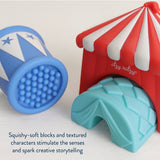 A children's sensory toy set on a light neutral background, featuring a red and white striped circus tent with a light blue textured entrance and a blue textured drum with white star accents. Text at the bottom reads, 'Squishy-soft blocks and textured characters stimulate the senses and spark creative storytelling.