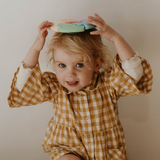 Child wearing a checkered dress with a colorful object on their head against a plain background