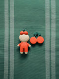A small decorative figurine of a child in an orange outfit with red cherries on the head, placed next to a pair of matching orange and green cherry-shaped accessories on a green striped fabric background.