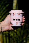 Hand holding a pink and white coffee cup with 'bbeáno' branding against a blurred green background