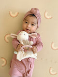 Baby in pink outfit holding a plush toy against a beige wall with decorative patterns