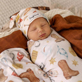 A sleeping newborn baby wrapped in a western-themed swaddle blanket and wearing a matching bonnet hat, resting on a brown blanket.