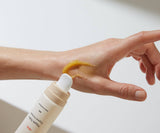 Hand applying a beige-colored cream from a bottle onto another hand against a light background
