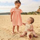 A toddler girl in a peach short-sleeve dress stands on a sandy beach looking down at a baby boy wearing a cream knit romper, who is sitting on the sand next to a large seashell.