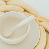 A white silicone baby spoon with the brand name 'renumer' printed on the handle, resting inside a matching white silicone bowl. The bowl is placed on a white plate, surrounded by sliced bananas on a clean white background.