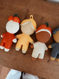 Four hand-painted wooden toy figurines with hanging loops, arranged on a rustic wooden table. The figures have minimalist faces and wear colorful outfits in orange, yellow, brown, and blue.