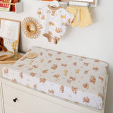 A baby changing pad covered in a white fitted sheet with a repeating teddy bear print, styled in a nursery setting with a matching baby outfit hanging above.