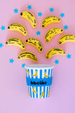 A blue and white striped paper cup with yellow banana prints and a blue band reading 'bbciño', surrounded by yellow banana-shaped decorations on a pink background with blue star cutouts.