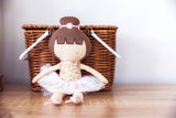 Handmade doll with brown hair and a floral dress sitting in front of a wicker basket on a wooden floor.