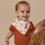 A smiling baby wearing a white bandana bib with a western print of boots, stars, and a guitar, styled with a rust-brown short-sleeve bodysuit. The baby is posed next to a brown cowboy hat and a bale of hay against a solid beige studio background.