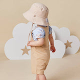 A baby wearing a beige sun hat, beige shortalls, and a light blue patterned t-shirt, standing in front of white cloud-shaped props with star cutouts on a beige background.