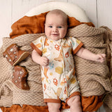 A baby lying on a cozy knit blanket and rust-colored fabric, wearing a short-sleeve one-piece romper with a western-themed print. A pair of brown cowboy boots with star details rests beside the baby.