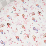 A close-up of a white crib bedding sheet or cover with a repeating watercolor print of mermaids, coral, starfish, and small sea creatures in pink, purple, and blue tones.