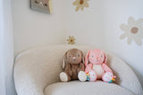 Two plush toys, a brown rabbit and a pink bear, sitting on a cushion against a wall with floral patterns.