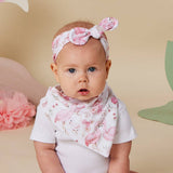 A baby wearing a white ribbed bodysuit, a pink flamingo-print triangular bib, and a matching pink flamingo-print bow headband, posed against a warm beige background with soft decorative props.