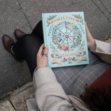 Person holding a book titled 'The Wheel of the Year' on a sidewalk.