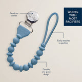 Blue pacifier clip with text highlighting features on a beige background