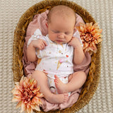 A sleeping newborn baby wearing a white ice cream-themed bodysuit, lying inside a woven oval basket lined with pink fabric and decorated with peach dahlia flowers, photographed from above on a textured cream blanket.