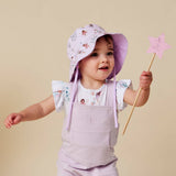 A toddler wearing a lavender sun hat with a floral print and matching lavender bib over a white t-shirt, holding a pink star wand against a solid beige background.