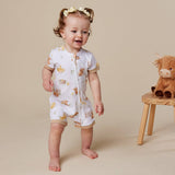 A toddler girl wearing a white short-sleeve footless romper with a yellow and brown animal print, standing barefoot on a beige studio floor next to a small wooden stool with a plush toy.