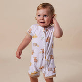 A toddler wearing a short-sleeve zip-front baby romper with a teddy bear print, standing against a plain beige studio background.