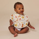 A baby sitting on a beige studio background wearing a white bib with a western-themed print of boots, hats, lasso, guitar, and stars.