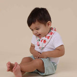 Baby sitting on a beige background wearing a white shirt with a floral bib and light green shorts.