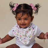 A baby wearing a white ribbed bodysuit and a white floral bandana bib, with dark hair styled in pigtails with pink bows, sitting against a solid beige background.