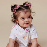 A smiling toddler with two buns of hair decorated with pink bows wears a white bib with a pink and multicolor ice cream cone pattern, sitting against a solid beige background.