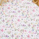 A close-up of a white fabric, likely a crib bumper or mattress protector, featuring a delicate watercolor-style floral and botanical print in pink, purple, blue, yellow, and green. The fabric is draped over a bassinet, with the wooden slats and a woven wicker basket edge visible in the background.