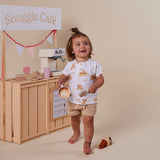 A toddler stands barefoot in front of a wooden play cafe setup, wearing a white short-sleeve top with a light brown bear and cake print, paired with beige shorts. The child holds a small toy coffee cup and looks to the side with a happy expression.