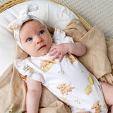 A baby lying on a beige blanket in a woven basket, wearing a white short-sleeve romper with a print of orange kittens and yellow fruits, along with a matching white headband.