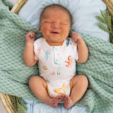 A newborn baby wearing a white short-sleeve bodysuit with a colorful sea-creature print, lying on their back in a wicker bassinet with a green knit blanket.
