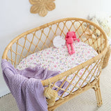 A natural rattan baby bassinet with a floral-patterned mattress, a pink plush monkey toy, and a purple knit blanket draped over the side, set against a white wall in a nursery setting.